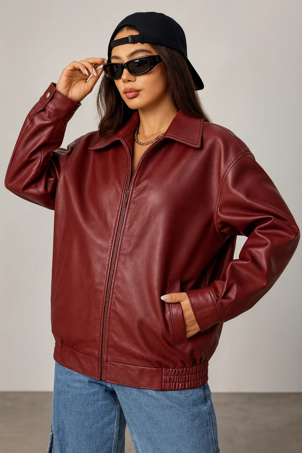 Woman in oversized maroon leather jacket, blue jeans, sunglasses, and black cap, urban style